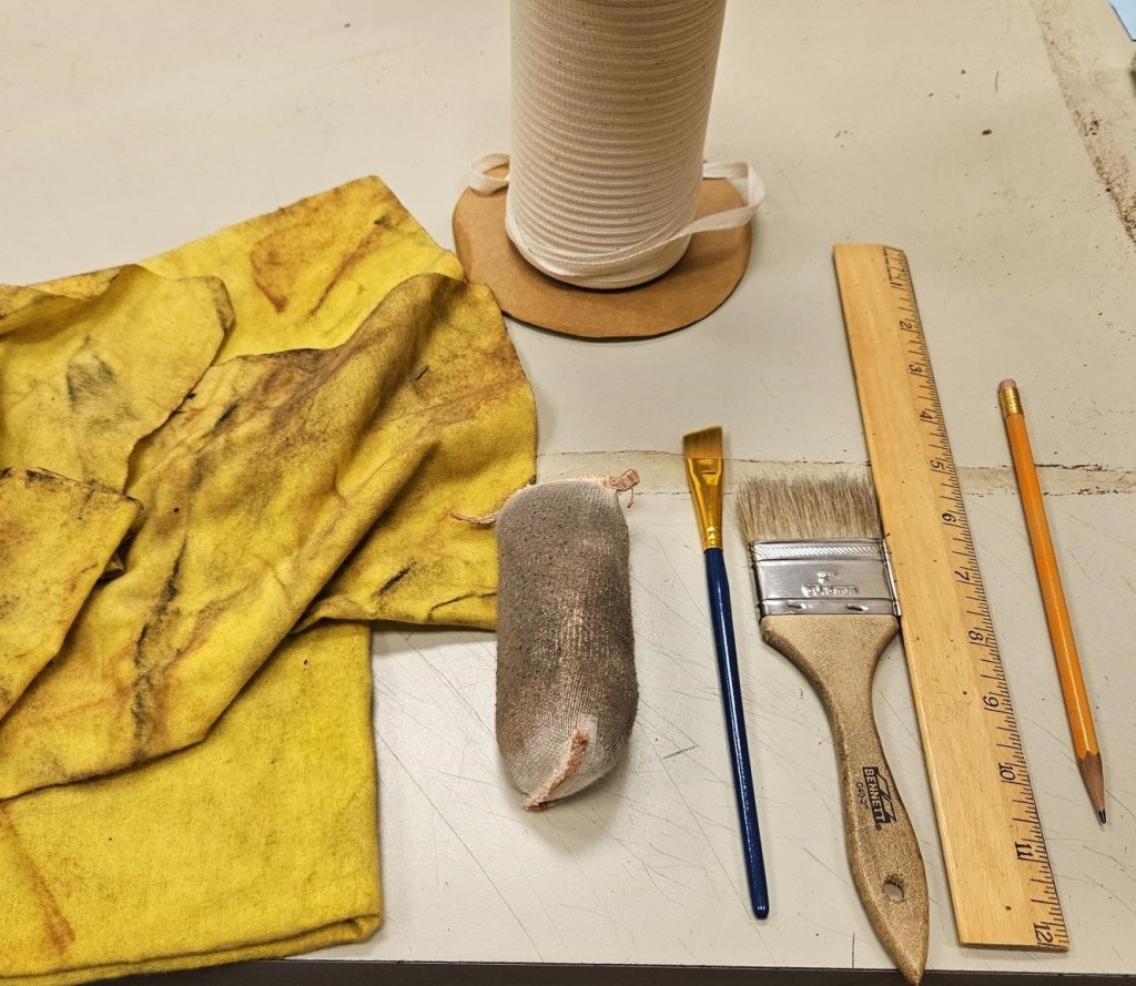 Photograph of cleaning supplies. There are dirty yellow dust cloths, a roll of white cotton twill tape, a dirty document cleaning pad, a small paint brush, a larger paint brush, a wooden ruler, and a pencil.
