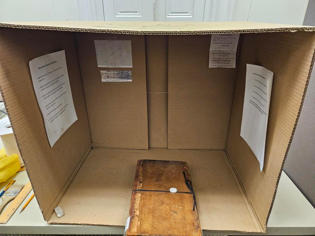 A photograph of a box on its side with a few pieces of paper taped on the inside. A old book sits on the inside.