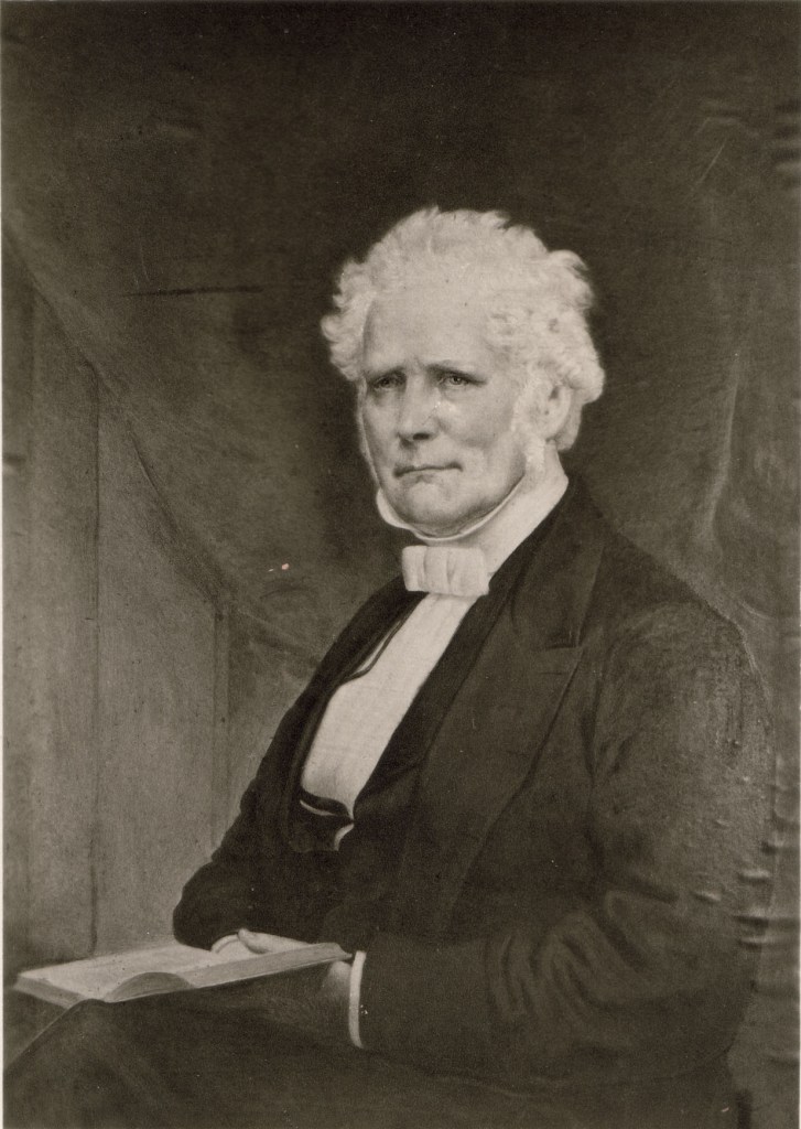 A black and white portrait of a gentleman with a head of white hair. He is sitting with his body positioned to the left. He wears a black suit with vest and white shirt with a high collar and white bowtie. He holds an open book in his lap.